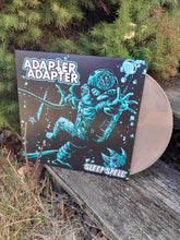 Charger l'image dans la galerie, Adapter Adapter 'Sleep Spell' vinyl record album shown outdoors on a weathered wooden surface with pine foliage in the background. The album cover is black with a glowing cyan creature illustration, and a translucent pink vinyl record is partially visible behind it.