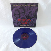 Charger l'image dans la galerie, Brown Acid - The Fifth Trip vinyl record with its sleeve, displayed on a white textured fabric background. The record is a distinctive translucent dark blue, and the sleeve features a purple-toned graphic collage with red and white text.