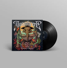 Charger l'image dans la galerie, A vinyl record partially pulled from its square album sleeve, shown on a light gray studio background. The cover art features a dark bird skull in a beaked mask with green glowing accents, surrounded by red, orange, and white decorative elements.