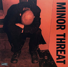 Load image into Gallery viewer, Album cover art for Minor Threat, featuring a crouched figure in black clothing with their head resting on their knees in a dim, orange-toned interior. The band name 'MINOR THREAT' is printed vertically in black on the right side, and a small 'DISCHORD' logo appears in the bottom-left corner.