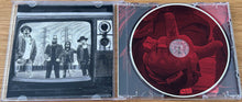 Cargar imagen en el visor de la galería, CD case with black and white illustration of a band on one side and red design on the other, on a wooden surface.