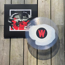 Cargar imagen en el visor de la galería, Vinyl record with a logo on a wooden surface next to a black cover with red and white design.