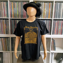 Load image into Gallery viewer, A black King Buffalo t-shirt on a mannequin, featuring a gold graphic design and the band's logo, with a white shelving unit full of vinyl records in the background.