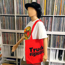 Load image into Gallery viewer, A red canvas tote bag with black 'Truck fighters' text displayed on a mannequin torso, set against a background of record store shelves filled with vinyl records.