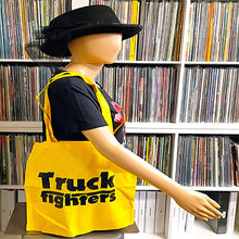 Load image into Gallery viewer, A bright yellow canvas tote bag with black text reading 'Truck fighters' is displayed on a mannequin torso in a record store, with shelves of vinyl records in the background.